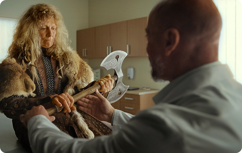 A hand specialist examines a patient in Viking-style furs holding a double-headed axe in a medical office.
